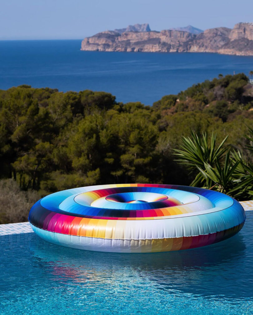 A colorful Felipe Pantone inflatable pool lounge floats in the pool, with a distant view of the ocean and mountainous coastline.