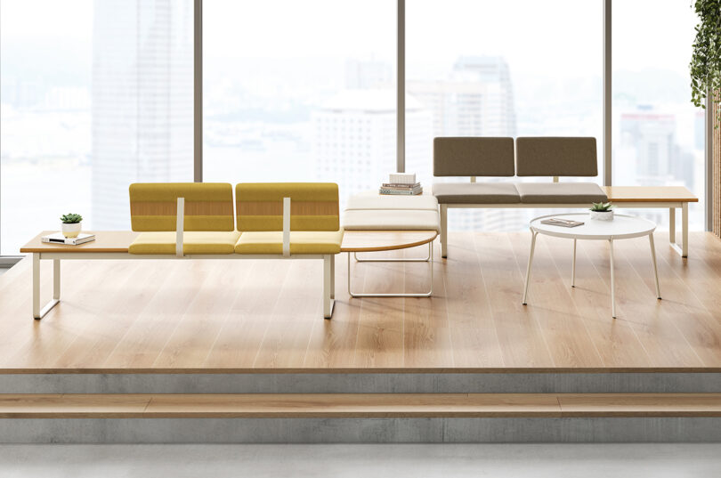 Modern office lobby with yellow and brown modular sofas, wooden tables, and large windows overlooking a cityscape.