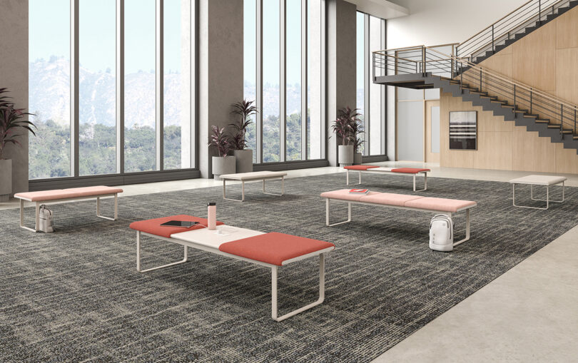 Modern office lounge with large windows showcasing mountain views, featuring several modular benches with red cushions and small coffee tables.