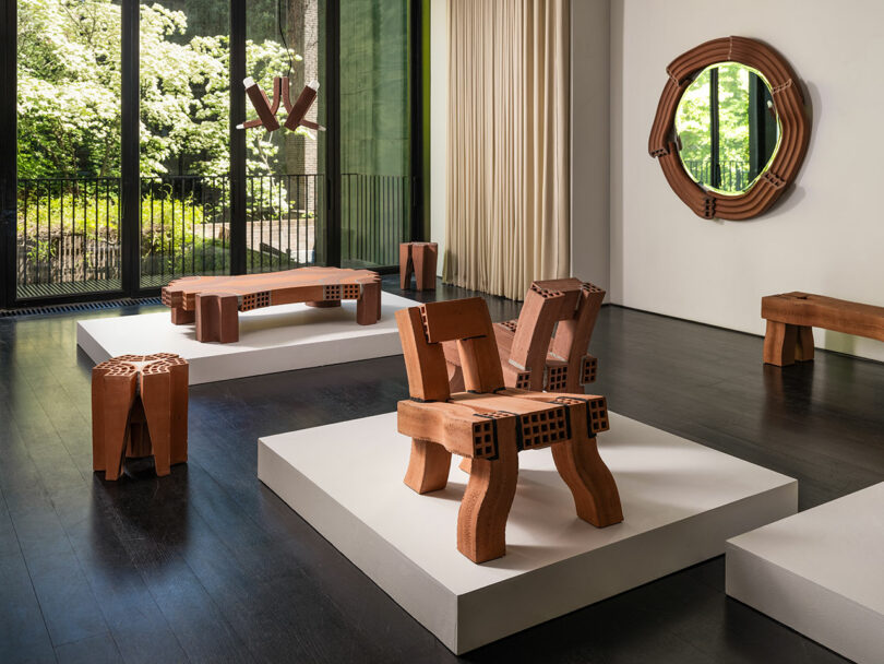 Uma galeria de arte moderna apresenta móveis de madeira, incluindo cadeiras, mesa e espelho com desenhos geométricos, expostos em plataformas com grandes janelas e árvores visíveis do lado de fora.