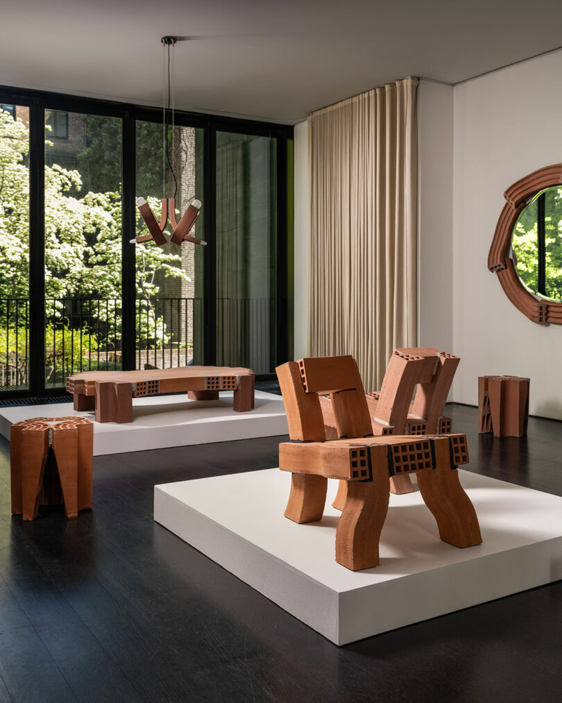 A modern art gallery features unique wooden furniture pieces on white platforms, large windows with a garden view, and light-colored curtains.