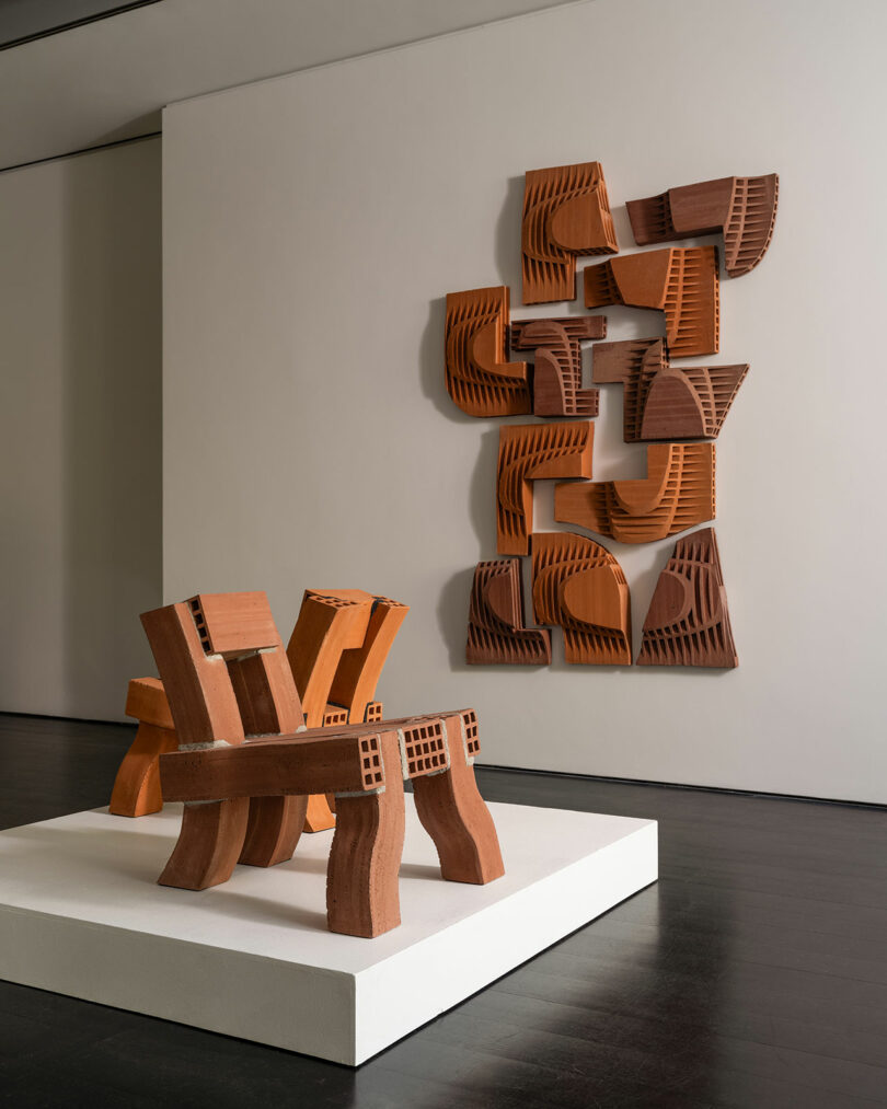 Uma instalação de arte moderna com esculturas de madeira dispostas geometricamente. Uma peça montada na parede composta por formas interligadas é exibida atrás de uma escultura abstrata de banco de madeira.