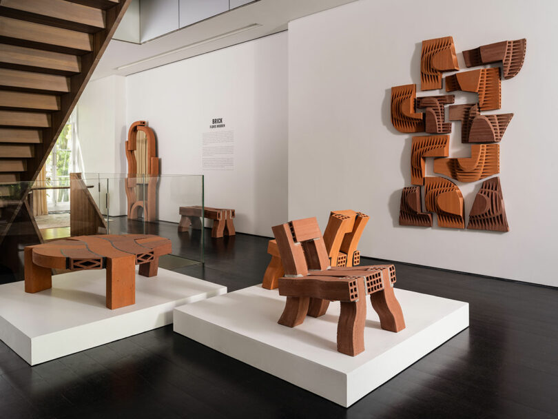 Uma exposição de arte moderna com móveis abstratos de madeira e arte nas paredes exibida sob uma escada em uma galeria.