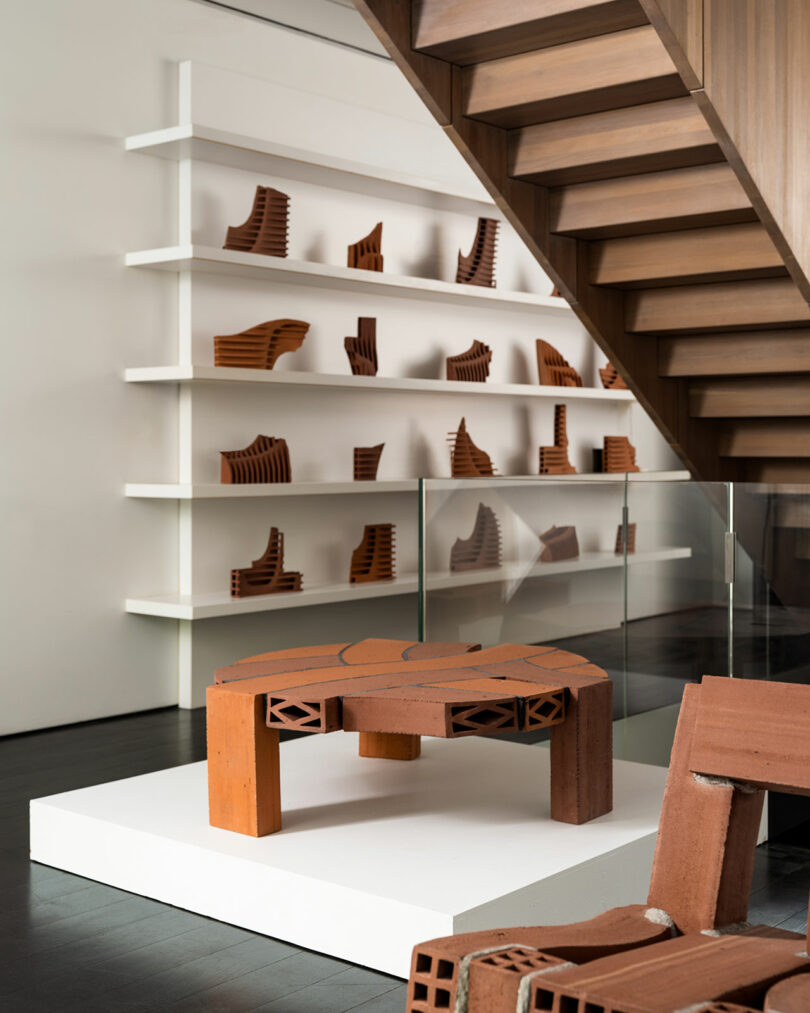 A modern interior displays terracotta sculptures and structures on white shelves by a wooden staircase. A showcase with glass barriers features a prominent terracotta piece on a white platform.