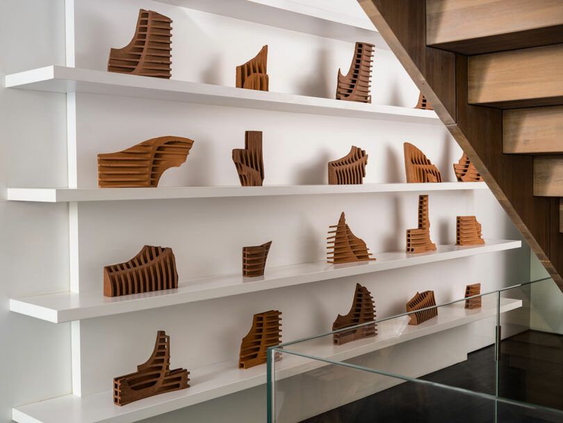 A display of various wooden architectural model sculptures on white shelves, positioned against a wall under a wooden staircase.