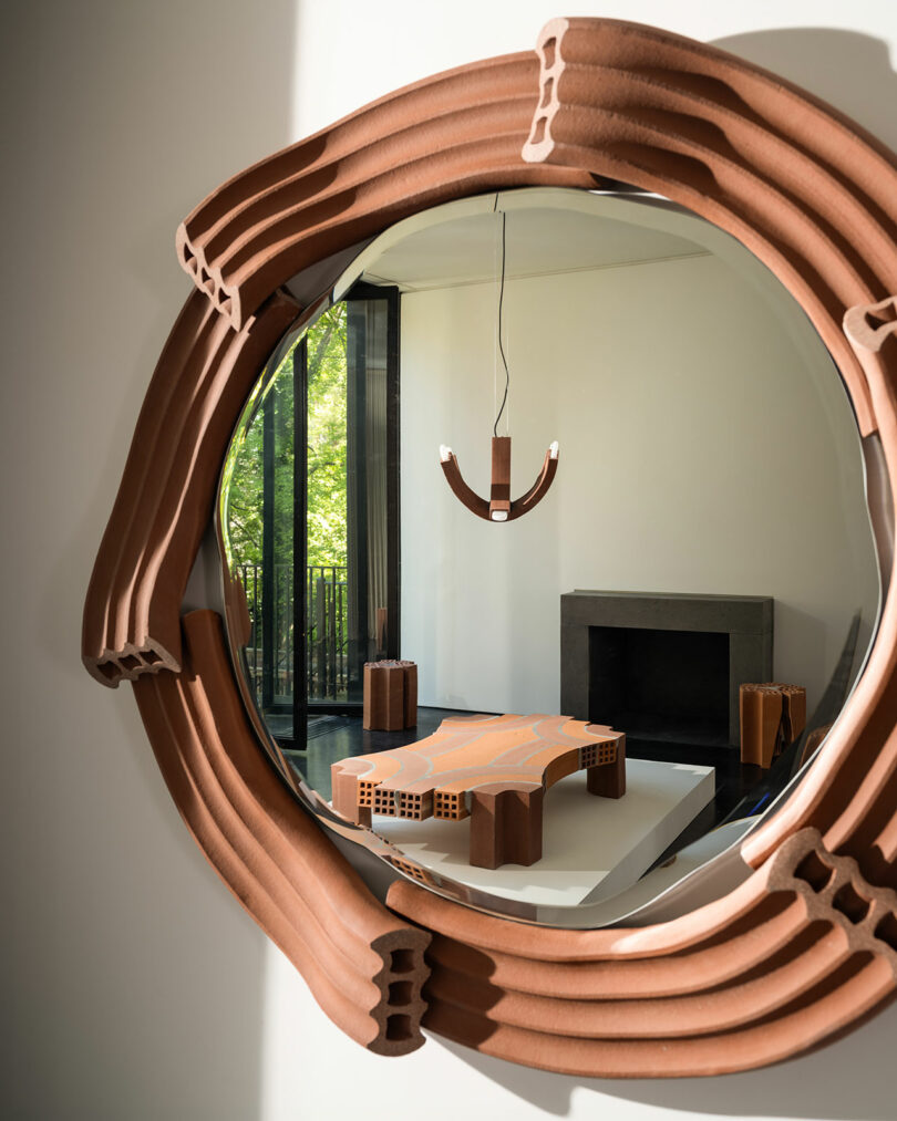 Uma sala de estar moderna com um grande espelho circular emoldurado por um material tipo terracota, refletindo uma mesa exclusiva e uma luminária pendente. Há uma lareira e grandes janelas ao fundo.