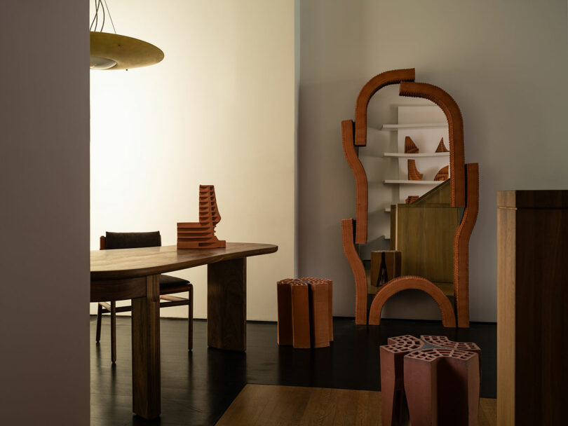 Quarto com móveis modernos de madeira, incluindo mesa, cadeira e estantes de formato exclusivo. Esculturas de cerâmica e objetos decorativos são colocados no chão e nas prateleiras.