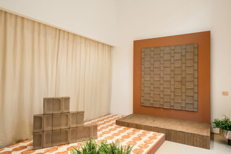 A minimalist room featuring a patterned floor, stacked brown blocks, a large textured wall panel, curtain backdrop, and scattered potted plants.