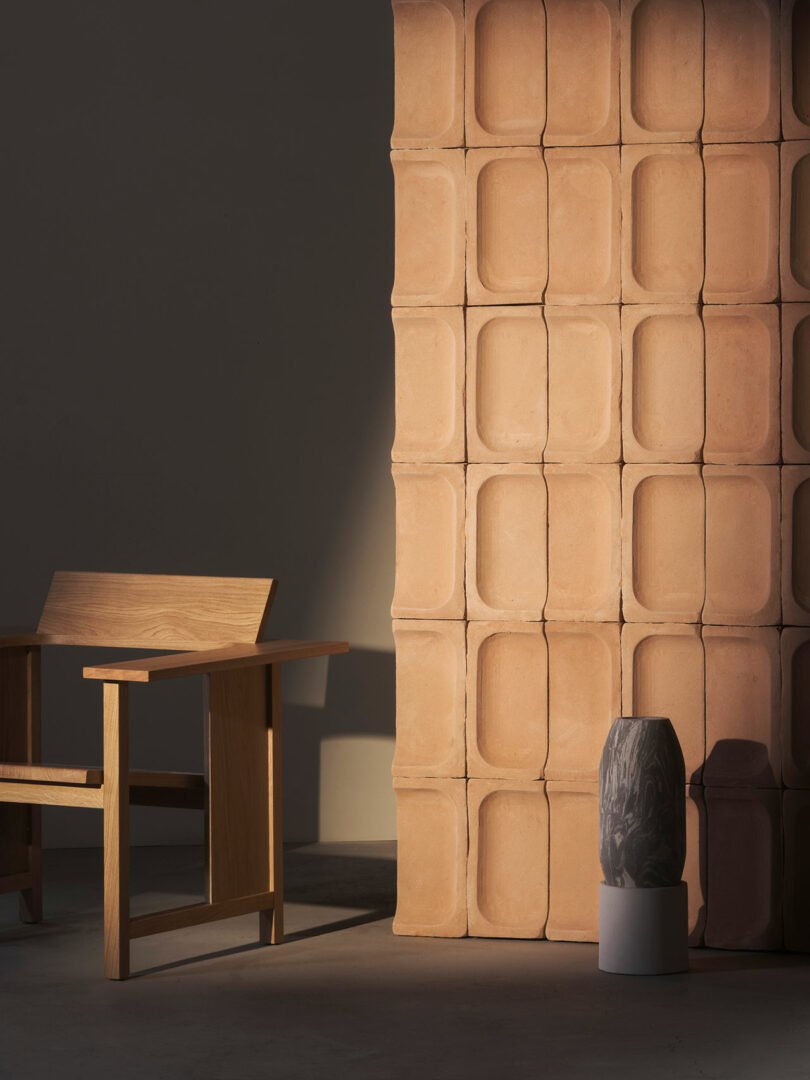 A minimalistic interior featuring a wooden chair, a textured clay tile wall, and two ceramic vases positioned on the floor.