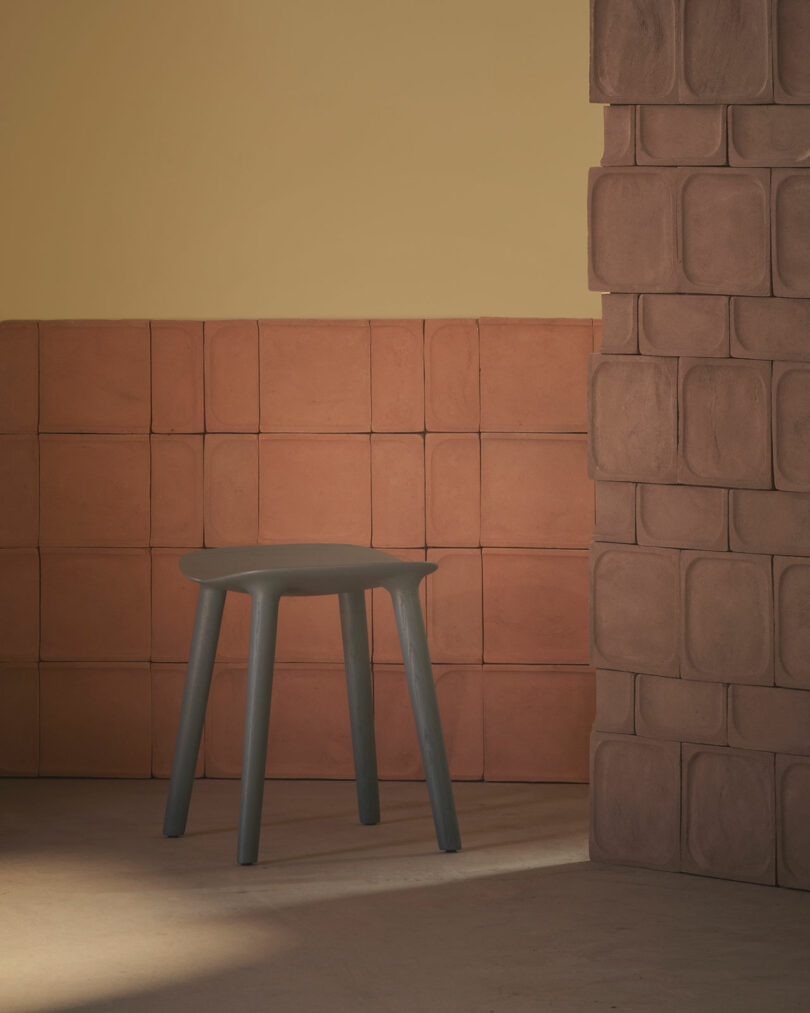 A dark gray stool stands against a backdrop of reddish-brown tiled walls in a room with warm lighting.