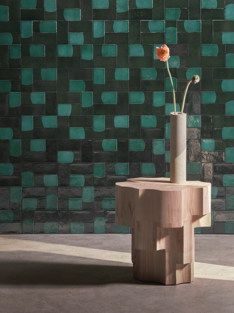 A wooden table holds a vase with two flowers in front of a green geometric tiled wall.