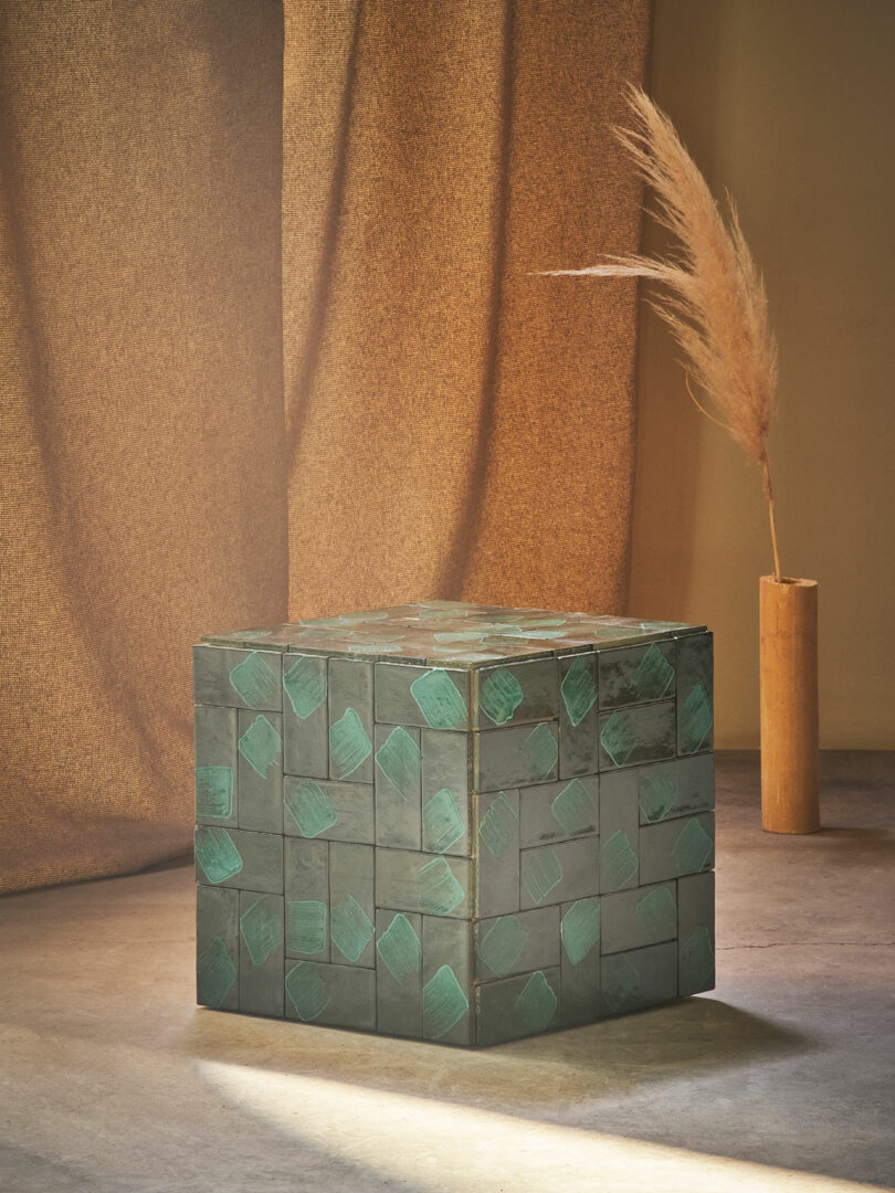 A geometric cube-shaped table with a green, mosaic pattern sits on a floor. Nearby, there is a tall, cylindrical vase with dried pampas grass against a brown, textured fabric backdrop.
