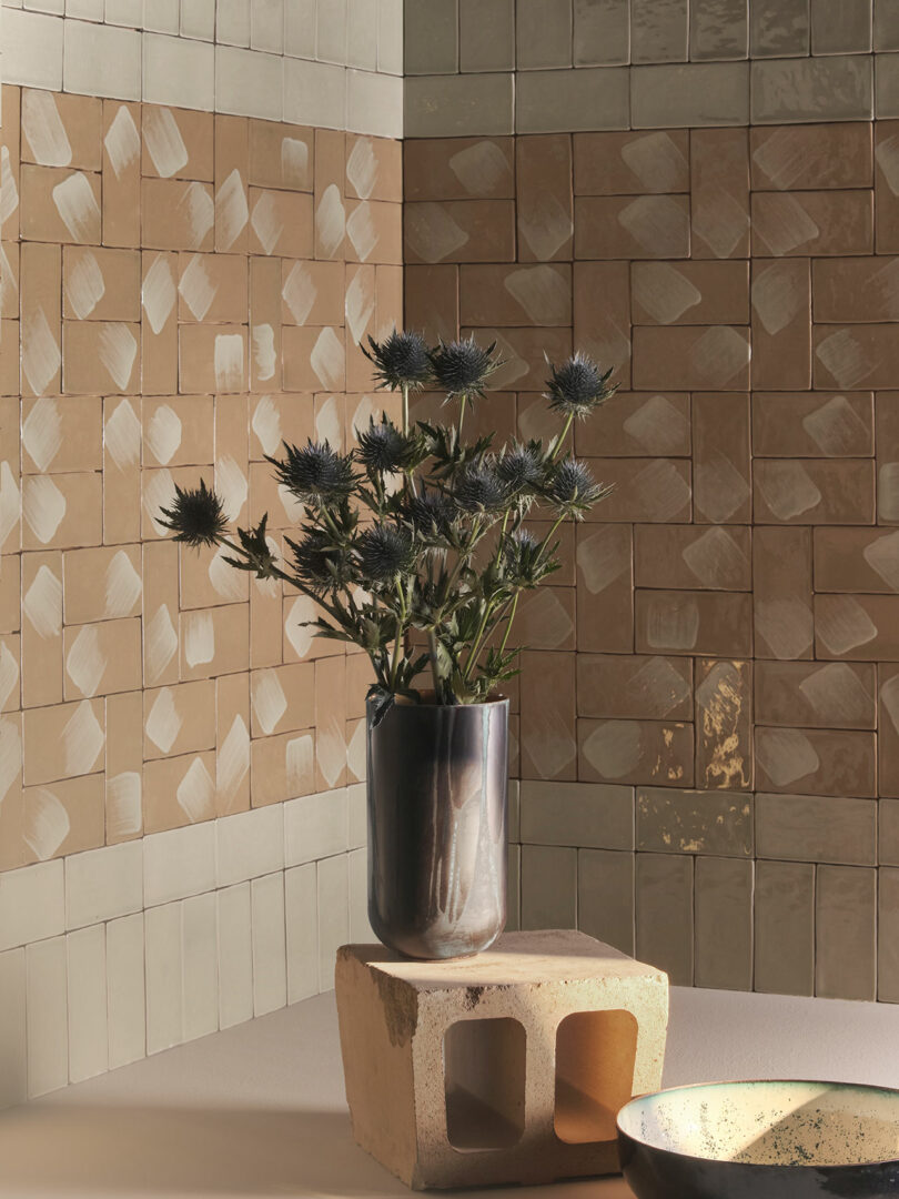 A ceramic vase with thistle flowers sits on a hollow block in front of a beige and tan tiled wall. A shallow circular dish is visible on the right.