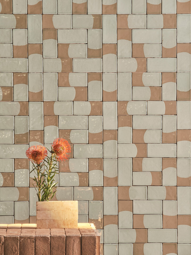 Rectangular tiles with a geometric pattern in beige and brown tones cover a wall. In the foreground, an arrangement of two orange flowers in a rectangular vase made of similar tile material.