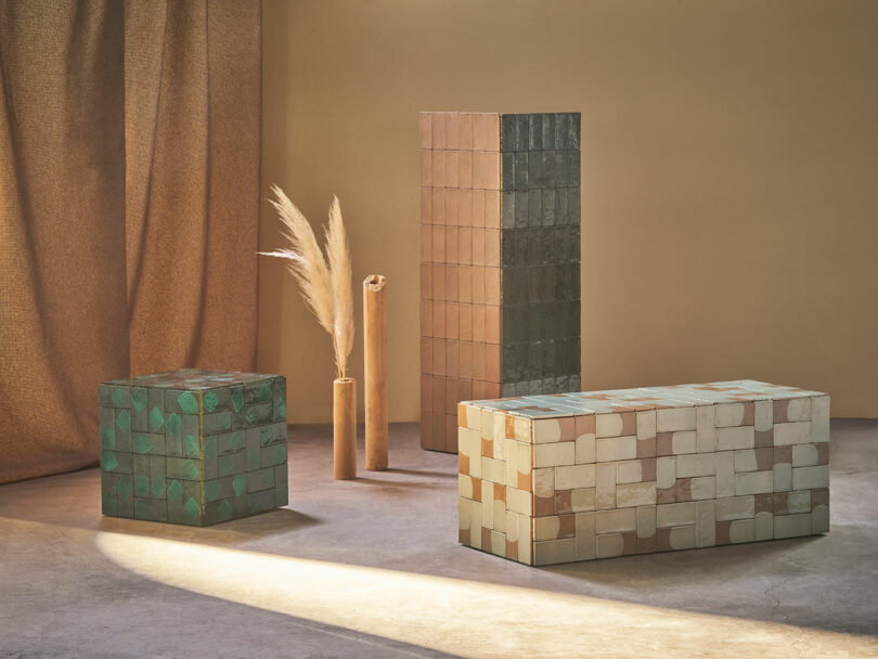 A room with four geometric, tile-covered furniture pieces in green and beige hues, arranged on a smooth floor. Dried pampas grass in a vase is placed among them. Soft light illuminates the setting.