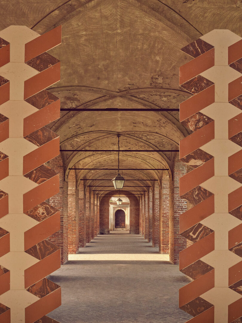 A long, vaulted corridor with brick arches and geometric patterns on the foreground columns. A hanging lamp is centered in the passageway.
