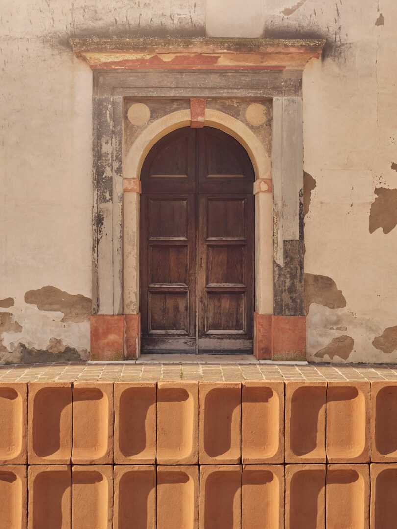 An old wooden arched door set in a weathered wall with faded paint and patches. In front of the door is a row of large, rectangular, orange tiles.