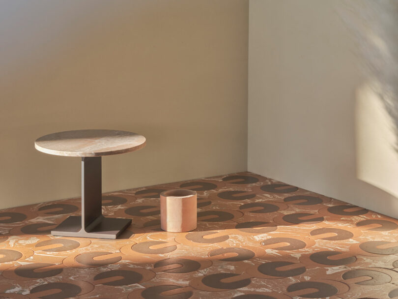 A minimalist room with a round table and a cylindrical stool on a patterned floor featuring large, curved shapes. The walls are smooth and light-colored, bathed in soft natural light.