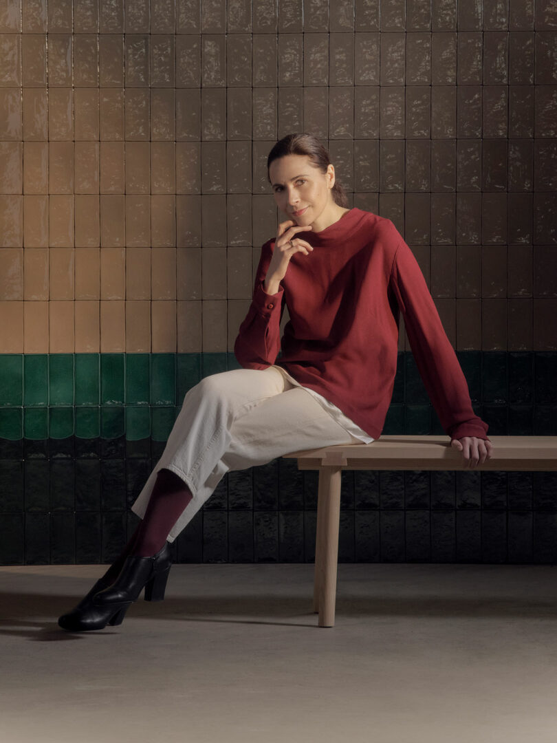 A woman sits on a wooden bench against a tiled background, wearing a red long-sleeve top, light pants, and black shoes. Her right hand is raised to her mouth while her left arm rests on the bench.