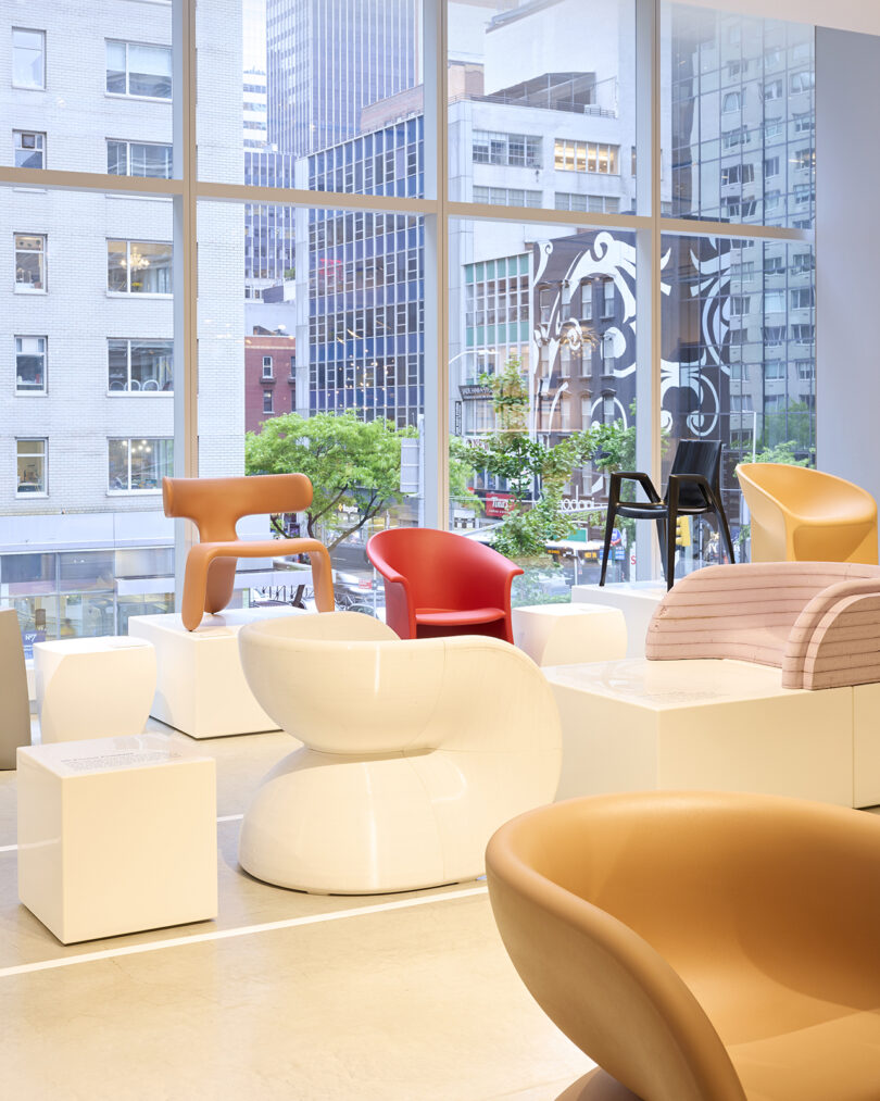 An exhibition room features modern designer chairs, including red, brown, and white pieces, displayed on white podiums in front of large windows with a cityscape view.