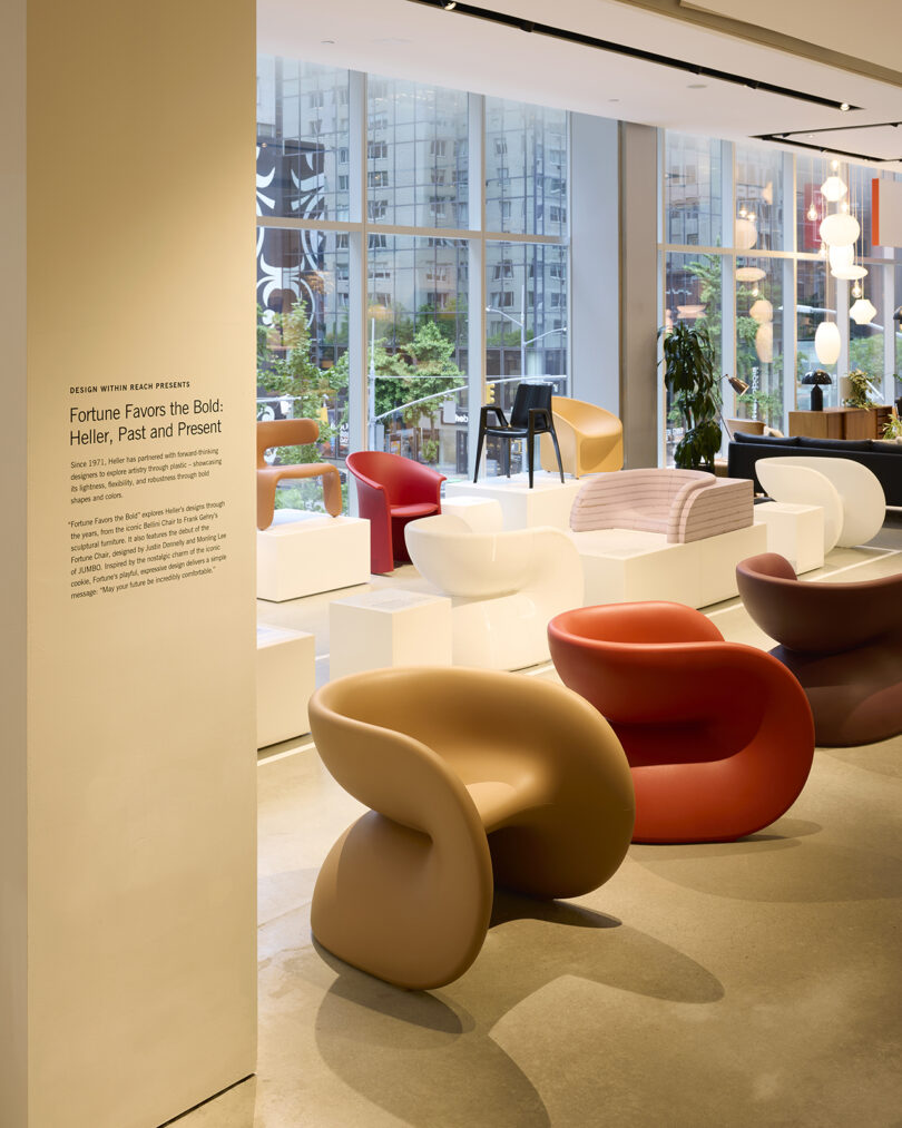 An exhibition room features modern designer chairs, including red, brown, and white pieces, displayed on white podiums in front of large windows with a cityscape view.