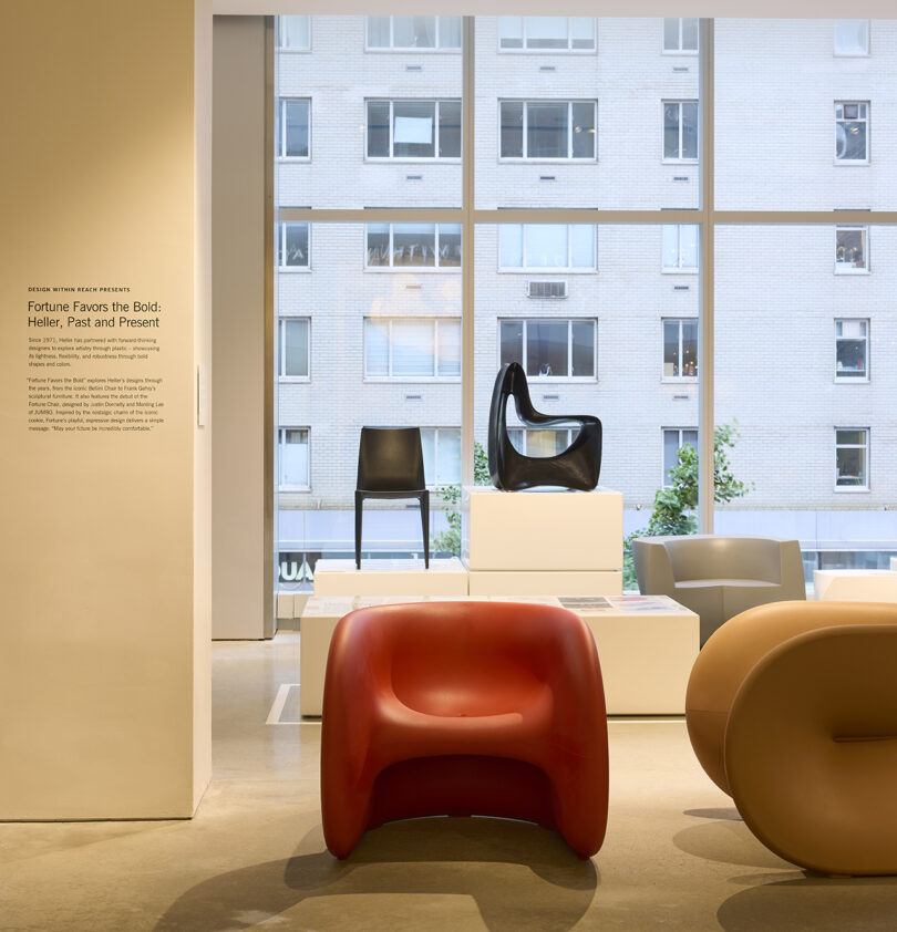 Uma sala de exposição apresenta cadeiras de design moderno, incluindo peças vermelhas, marrons e brancas, exibidas em pódios brancos em frente a grandes janelas com vista para a paisagem urbana.