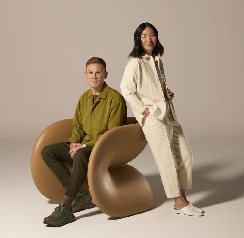 Two people pose for a photo; one is seated on a modern, curved chair in a green outfit, while the other stands beside them in a light-colored outfit. The background is neutral.