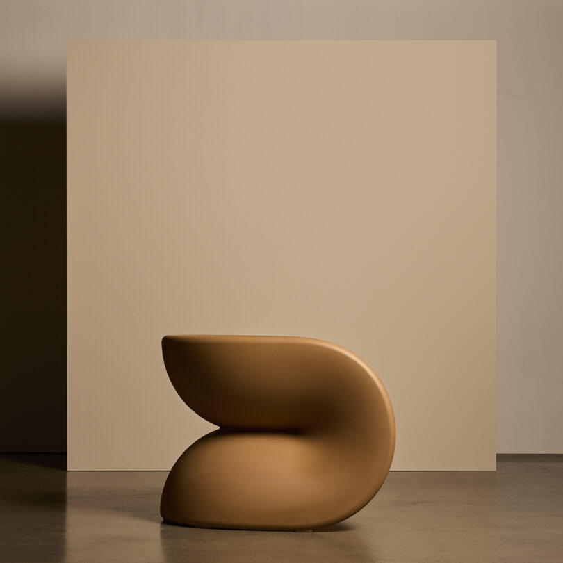 A modern, sculptural chair with smooth, curved lines in a beige color, placed in front of a matching beige backdrop.