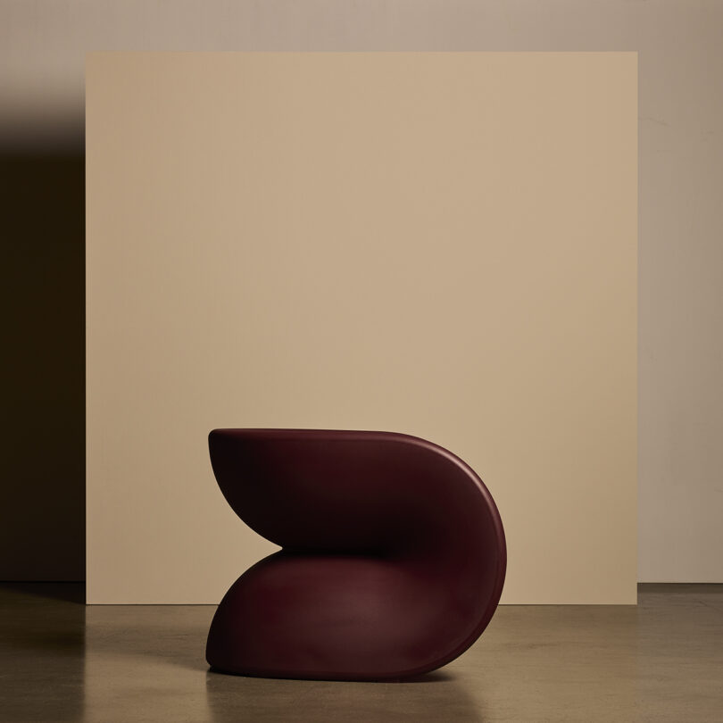 A modern, sculptural chair with smooth, curved lines in a maroon color, placed in front of a beige backdrop.