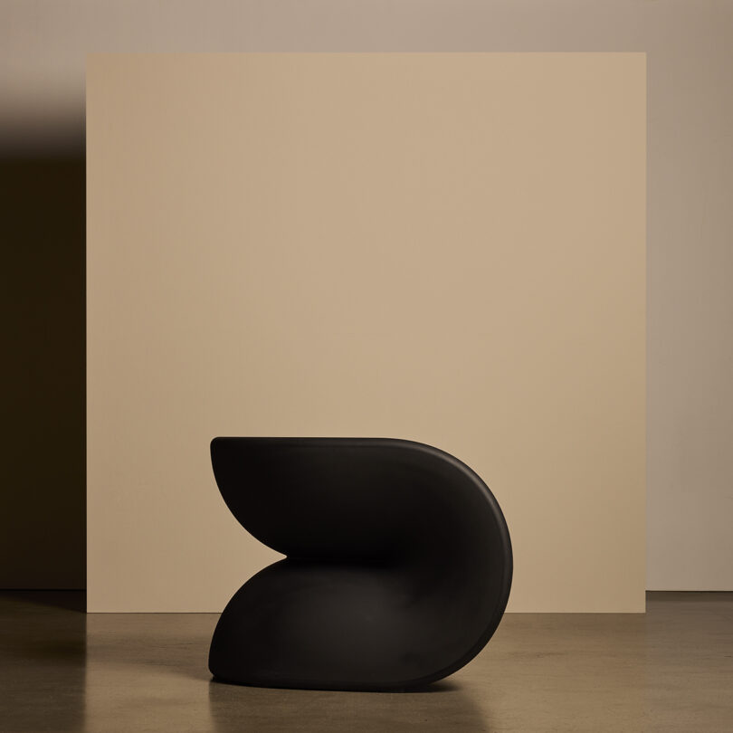 Uma cadeira moderna e escultural com linhas suaves e curvas em preto, colocada em frente a um fundo bege.