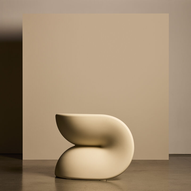 Uma cadeira moderna e escultural com linhas suaves e curvas em creme, colocada em frente a um fundo bege.