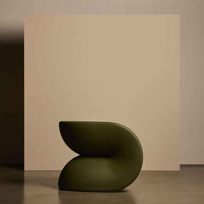 A modern, sculptural chair with smooth, curved lines in dark green, placed in front of a beige backdrop.