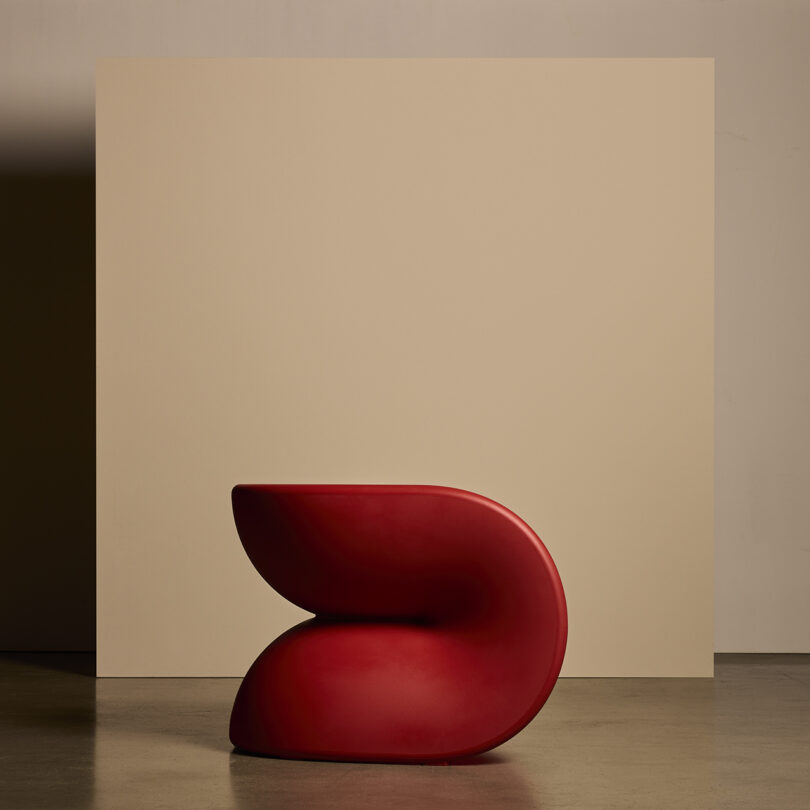 Uma cadeira moderna e escultural com linhas suaves e curvas em vermelho, colocada em frente a um fundo bege.