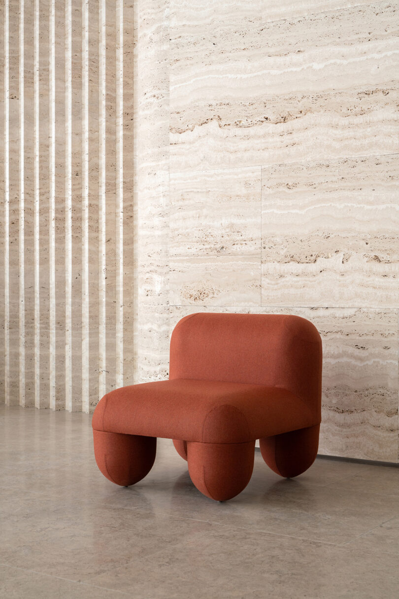 A modern red chair with a curved design positioned against a textured beige marble wall, with vertical stripe patterns to the side.