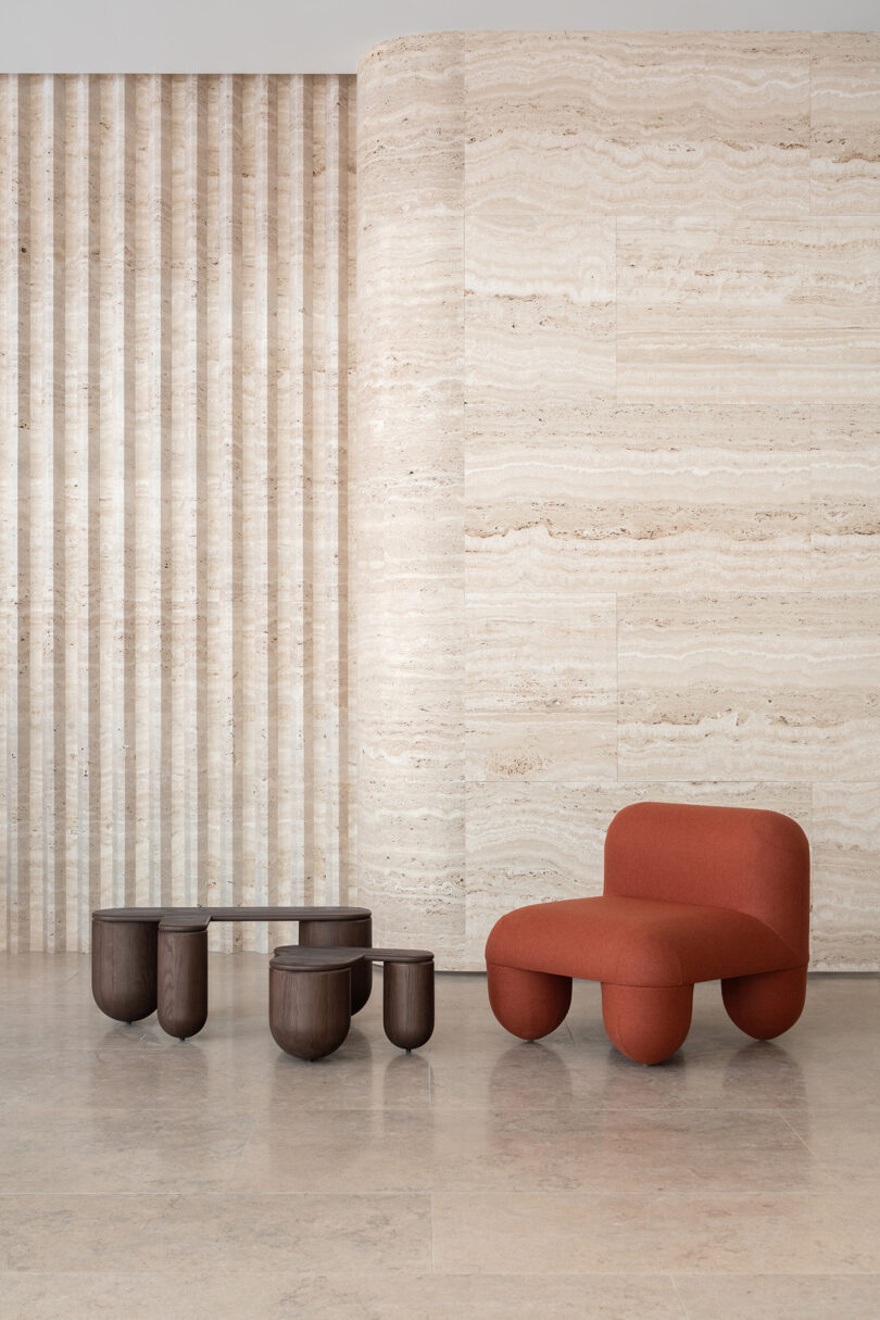 A modern red chair with a curved design and two dark wood coffee tables positioned against a textured beige marble wall, with vertical stripe patterns to the side.
