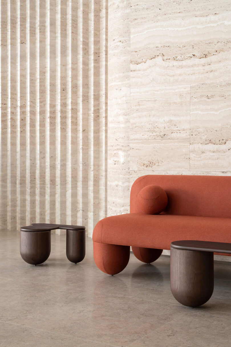 A modern red sofa with a curved design and two dark wood coffee tables positioned against a textured beige marble wall, with vertical stripe patterns to the side.