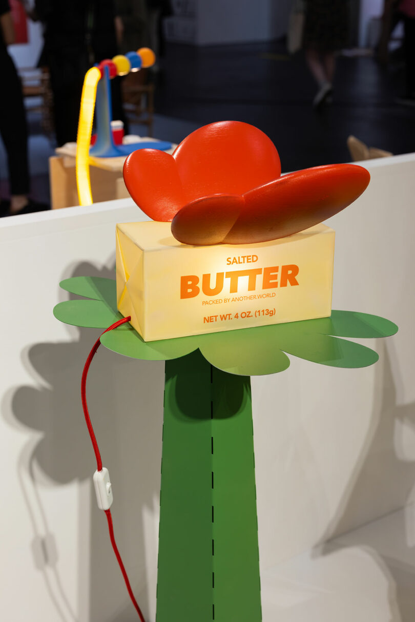 A large plastic butter sculpture light labeled "SALT BUTTER" and "PACKED BY ANOTHER WORLD" is displayed on top of a green pedestal with a red and yellow butterfly decoration attached, resembling an artistic launch pad ready to take off.