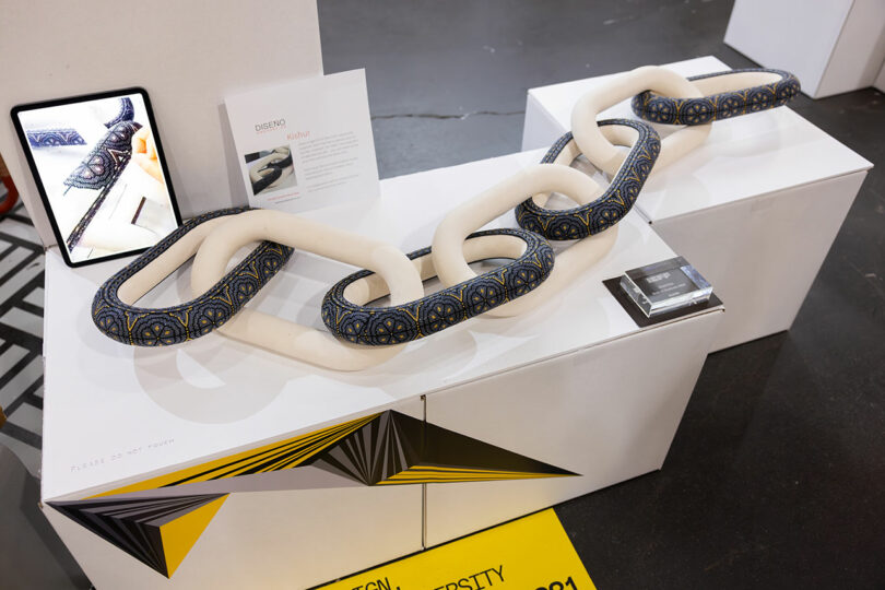A display on a white table features a snake-like chain sculpture, an iPad showing a detailed close-up, and an informational card. Yellow and black geometric shapes surround the setup, creating a dynamic launch pad for the artwork.