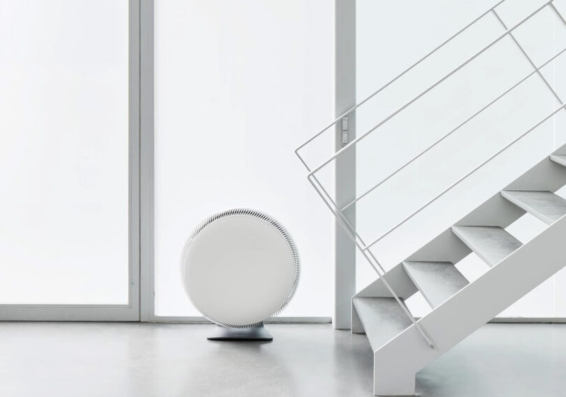 A round, white IQ Air Atem X sits near the base of a modern, white staircase in a minimalist room with large windows.
