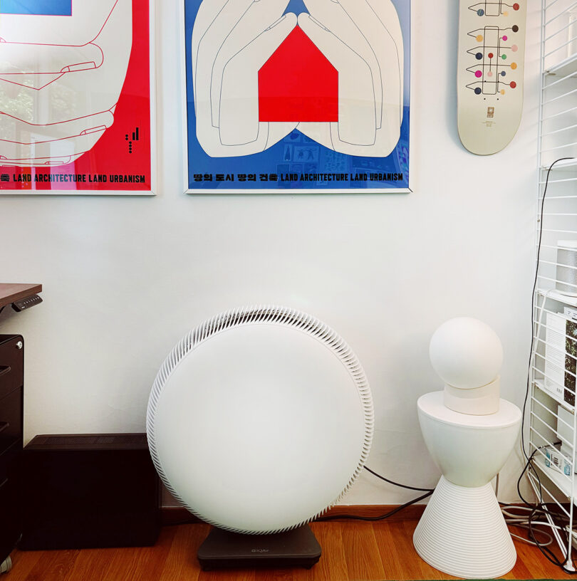 A modern home office featuring an IQ Air Atem X circular air purifier, a small white lamp on a round side table, and wall-mounted art posters and shelves.