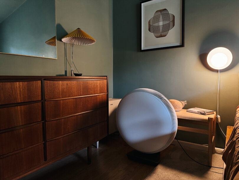 A cozy room with a wooden dresser, a framed geometric artwork, a bench with a book and lamp, and an IQ Air Atem X air purifier beside the large circular fan on the floor.