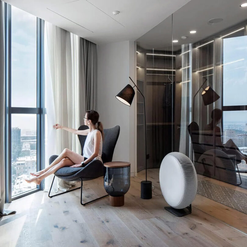 A person sits on a chair by a large window in a modern, minimalist room with wooden flooring and mirrored walls, looking out at a cityscape. A floor lamp and an IQ Air Atem X air purifier are nearby.