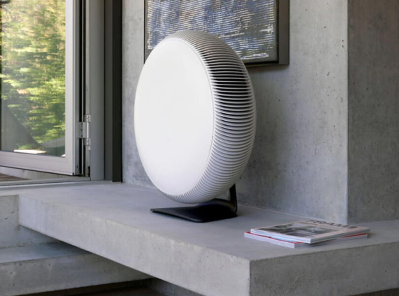 An IQ Air Atem X, white and round, rests on a concrete ledge next to a magazine in a modern room with large windows.