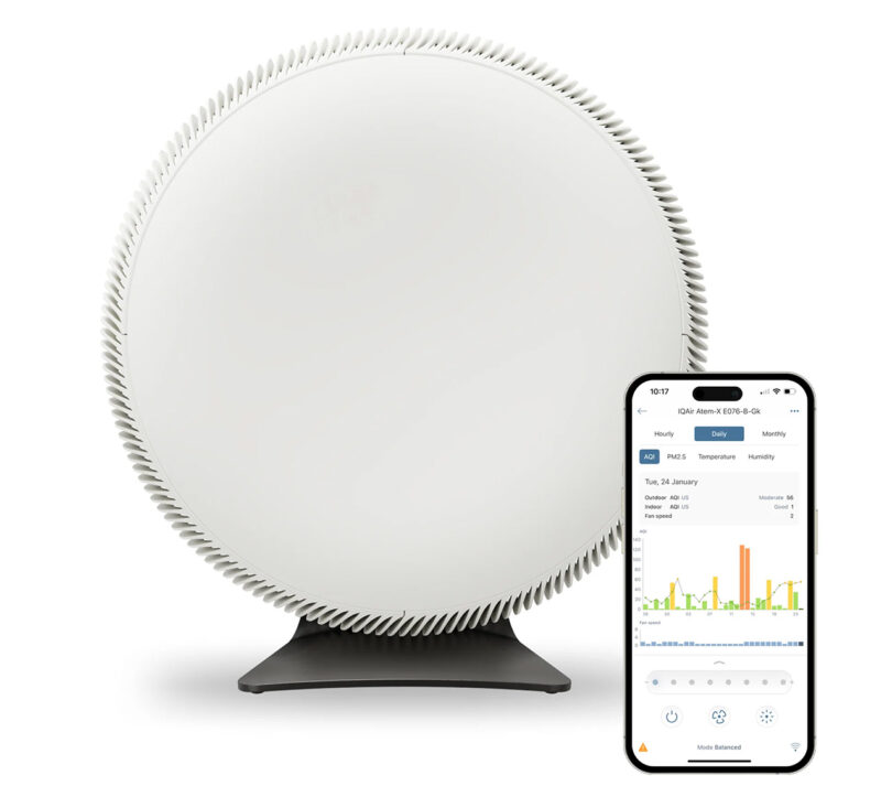 A white, circular IQ Air Atem X air purifier on a black stand is shown beside a smartphone displaying air quality data on its screen.