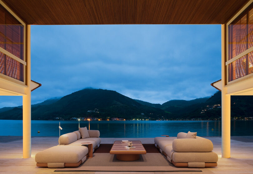 Modern open living space with beige modular sofas and a coffee table overlooks a serene lake and mountains under a cloudy evening sky.