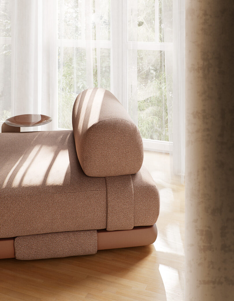 A modern beige sofa positioned near a window with sheer curtains, allowing natural light to flood the room. A small round side table is adjacent to the sofa.