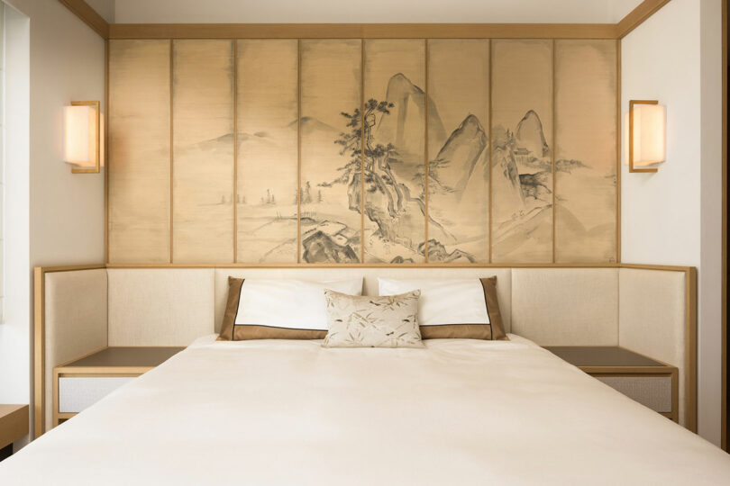 A minimalist bedroom with a large bed centered against a paneled wall featuring a traditional asian landscape painting, flanked by two wall-mounted lamps.