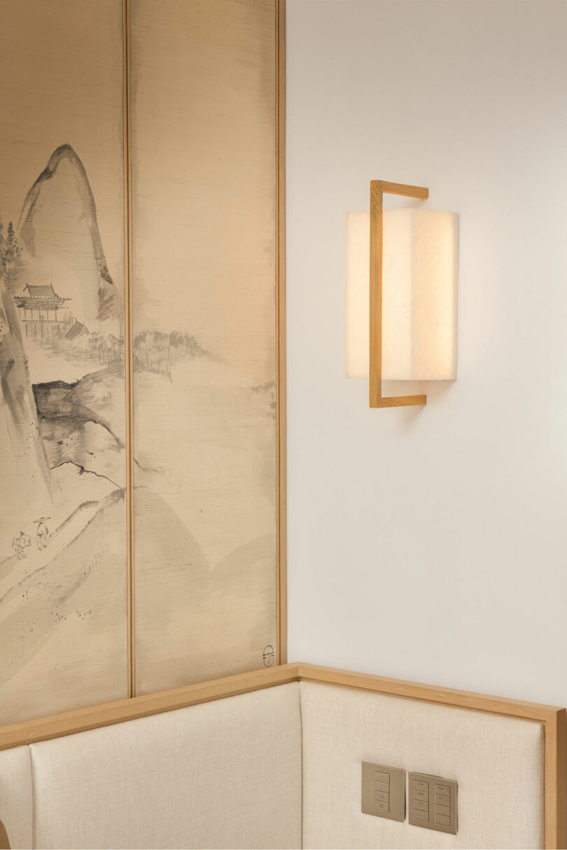 A modern wall lamp on a beige wall next to a tall, narrow painting of a traditional asian landscape, above a light-colored upholstered bench.