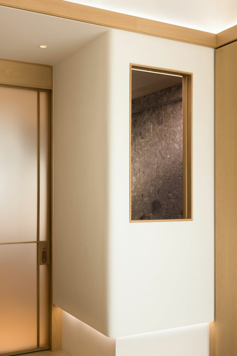 Minimalist interior with a cream-colored hallway featuring a wooden-framed rectangular window showing a textured brown wall.