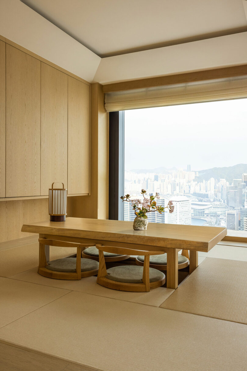 A minimalist japanese-style dining area with a low wooden table, floor cushions, and a large window overlooking a cityscape.